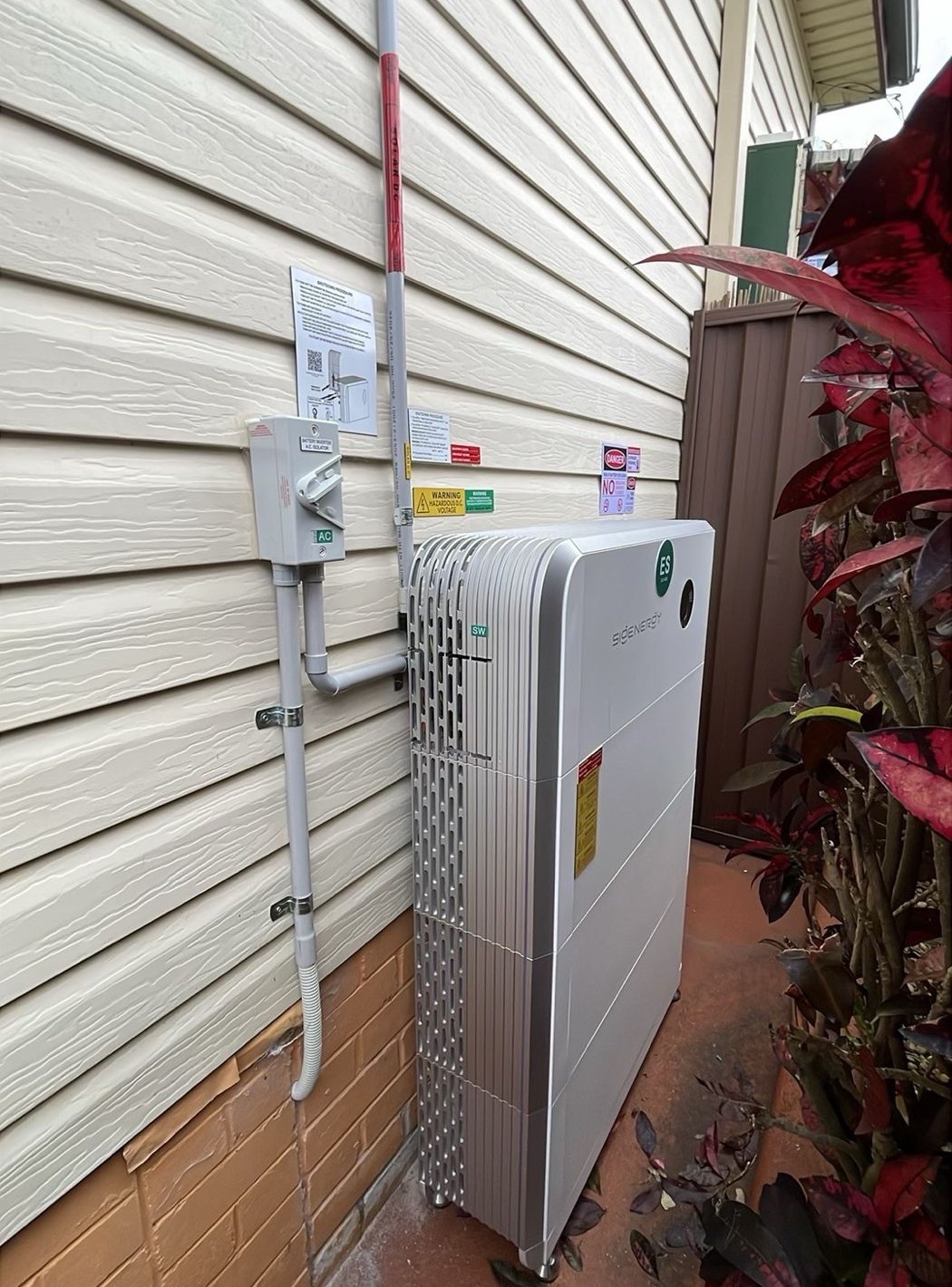 Sigenergy SigenStor with three battery modules on a side wall, AC isolator and labelled conduit beside it.
