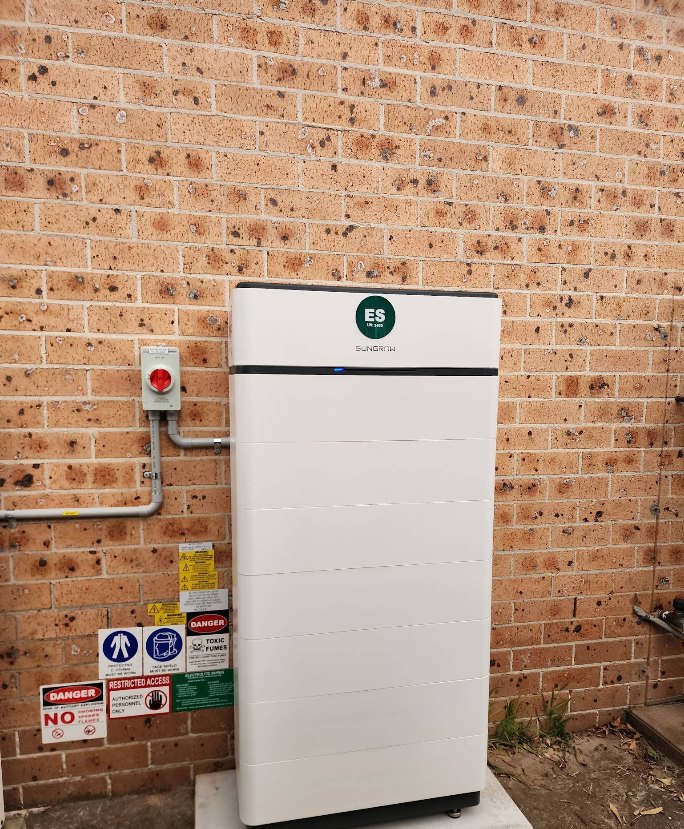 Stacked Sungrow battery installed against a brick wall with isolator and hazard signage.